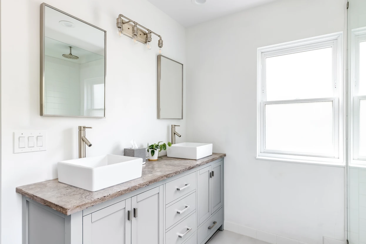 Une salle de bains élégante et rénovée avec des lavabos blancs, une vasque grise, un plan de travail en granit et du matériel en bronze, des robinets et de la lumière